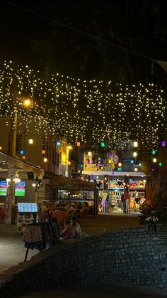 people are sitting on benches in the middle of an open area with lights strung overhead