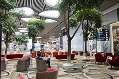 a large lobby with lots of red chairs and trees in the center, while people are sitting on couches