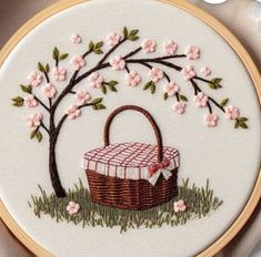 an embroidery project with a basket on the grass and cherry blossom trees in the background