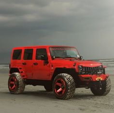 a red jeep parked on top of a sandy beach next to the ocean under a cloudy sky