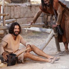 a man sitting on the ground next to another man holding a wooden stick in his hand