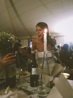 a woman sitting at a table with wine glasses in front of her and people standing around