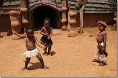 three young children are playing in front of an old hut with thatched roof and stone walls