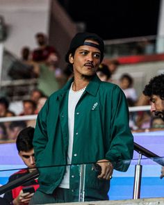 a man in a green jacket and hat standing next to a fence with people watching