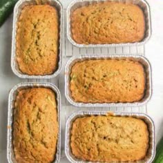 six baked zucchini breads sitting in tins on a white surface next to cucumbers