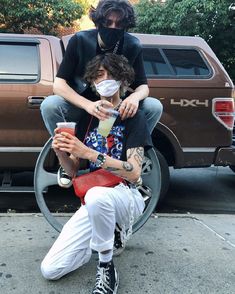 two people sitting on the back of a truck, one with a face mask and another holding a drink