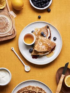 pancakes with blueberries, lemons and syrup on a yellow tablecloth next to bowls of fruit