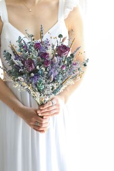 a woman in a white dress holding a bouquet of purple and green flowers on her arm