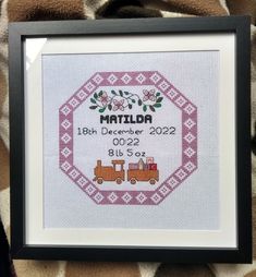 a cross - stitch birth announcement is displayed in a black frame on a patterned blanket