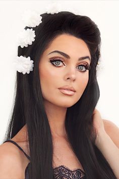 Jackie Wyers wearing a black wig with white flowers throughout the crown of the head. Her makeup is in the style of Priscilla Presley with thin dark brows, graphic liner, a tan and a frosty nude lip with a darker lip liner. Priscilla Presley 60s, Hair With Flowers, Mod Makeup, Look Disco