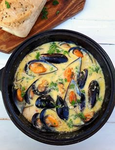 a bowl of mussels and bread on a white wooden table next to a cutting board