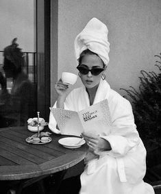 a woman in a bathrobe sitting at a table reading a book and drinking coffee