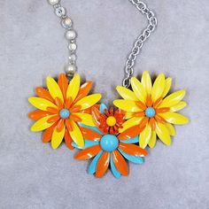 an orange, yellow and blue flower necklace is hanging from a chain on a gray surface