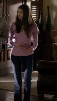 the young woman is standing in her living room wearing jeans and a pink shirt with gold accents