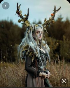 a woman with long white hair and horns standing in tall grass wearing a costume that has deer antlers on it's head