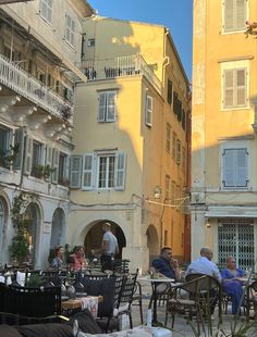 people are sitting at tables in an alleyway with yellow buildings behind them and one man is walking