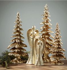 two gold angel figurines standing in front of some christmas trees on a wooden table