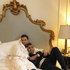 two men sitting on a bed with white sheets and pillows, one is holding his arm around the other's shoulder