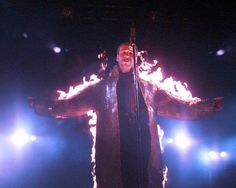 a man standing on stage with his arms open and fire in the air behind him