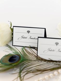 wedding place cards with peacock feathers and white roses on the side, sitting next to each other