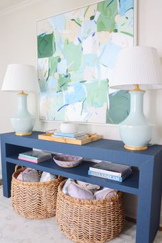 a blue table with two baskets underneath it and a painting on the wall behind it