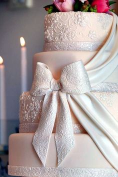 a three tiered wedding cake with white ribbon and pink flowers on top, sitting next to two lit candles