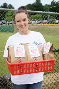 a woman is holding up some boxes in front of a chain link fence at a baseball field