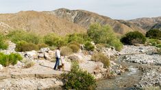 two people walking along a rocky riverbed in the desert with mountains in the background