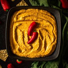 a square pan filled with yellow hummus next to crackers and red chili peppers