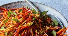 a bowl filled with carrots and greens on top of a blue table cloth next to a metal spoon