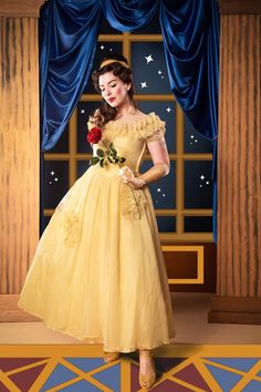 a woman in a yellow dress is holding a rose and posing for a photo with a blue curtain behind her