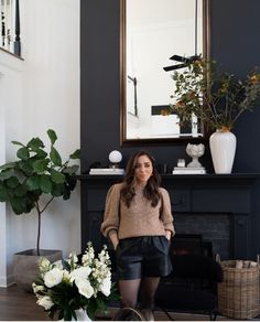 a woman standing in front of a fireplace with flowers on the mantle and potted plants