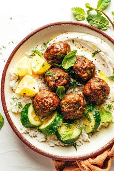 a bowl filled with meatballs and rice on top of a white table next to sliced cucumbers