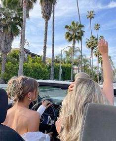 two women sitting in a convertible car with palm trees behind them