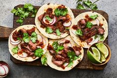 four steak tacos on a wooden cutting board with limes and cilantro