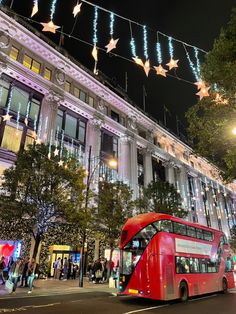 a red double decker bus parked in front of a tall building with christmas lights hanging from it's sides