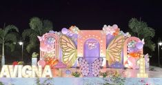 the entrance to an amusement park decorated with balloons and flowers at night time, surrounded by palm trees