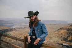 a man with long hair wearing a cowboy hat and jeans leaning on a wooden fence