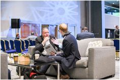 two men sitting on couches talking to each other in an office lobby with tables and chairs