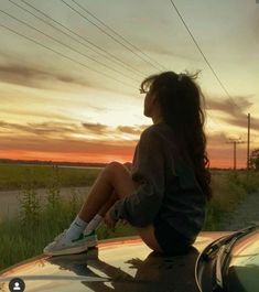 a woman sitting on top of a car next to a body of water at sunset