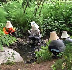 three people in hats are sitting on the ground near a stream and some bushes, while one person is looking at his cell phone