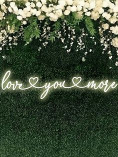 the words love you more are lit up in front of white flowers and greenery