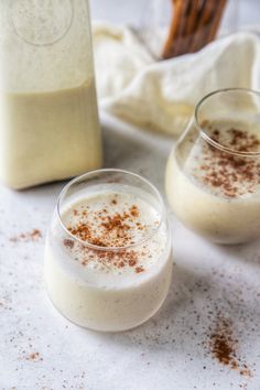 two glasses filled with milk next to a bottle and cinnamon stick on a white surface