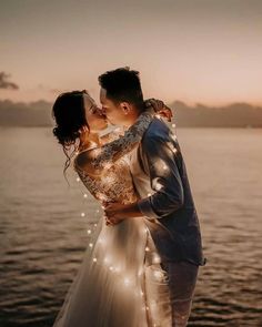 a bride and groom kissing in front of the water at sunset with lights on them