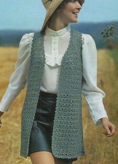 a woman wearing a crocheted vest and hat walking through a field with her hand in her pocket