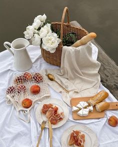 a picnic table with bread, fruit and cheeses on white cloth next to water