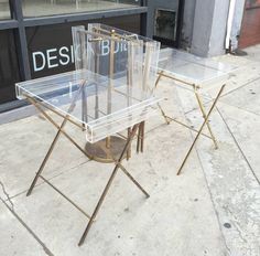 two clear tables sitting on the sidewalk in front of a storefront with designs written on it