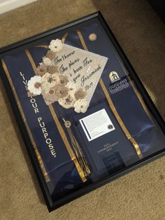 a graduation cap and flowers on display in a shadow box with gold ribbon around it