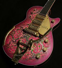 a pink and gold guitar sitting on top of a black surface with an intricate design