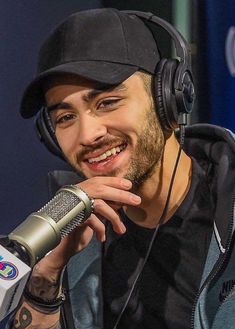 a smiling man with headphones on his ears and wearing a black cap, sits in front of a microphone